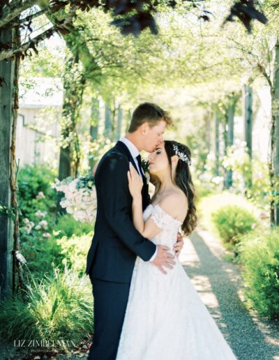 A bride and groom share a kiss on a path surrounded by greenery, with sunlight filtering through the trees.