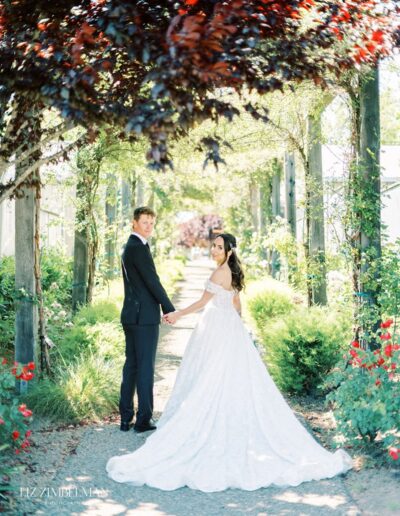 A bride and groom hold hands under a leafy pergola. The bride wears a white gown, and the groom is in a black suit. The path is lined with greenery and red flowers.