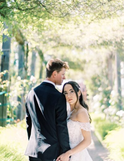 A couple in wedding attire stands outdoors on a sunny path, holding hands. The woman looks directly at the camera, while the man gazes to the side. Trees and greenery surround them.