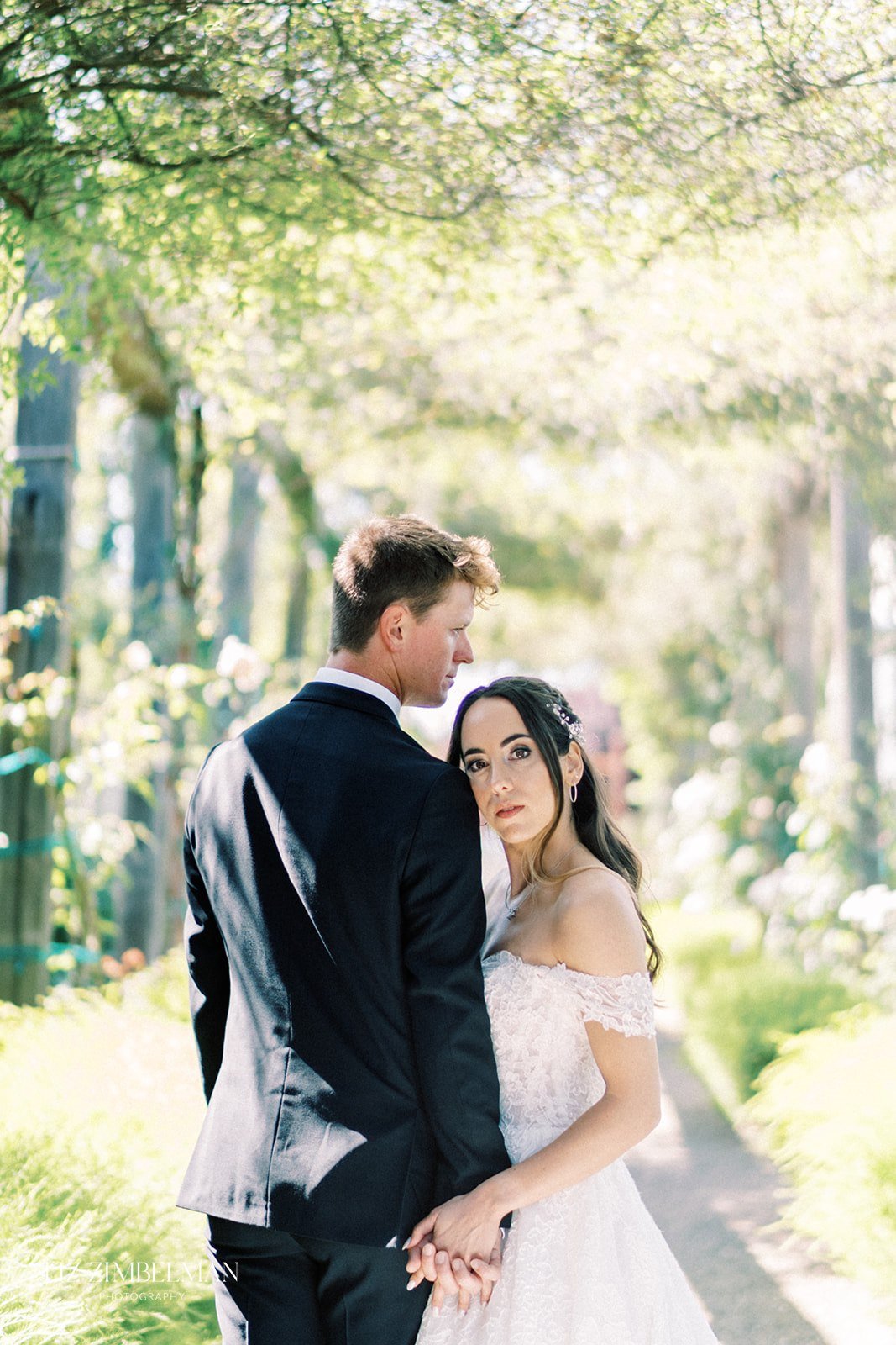 A couple in wedding attire stands outdoors on a sunny path, holding hands. The woman looks directly at the camera, while the man gazes to the side. Trees and greenery surround them.