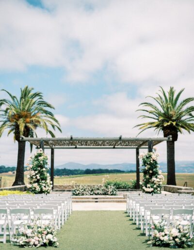 Outdoor wedding setup with white chairs facing a pergola adorned with flowers. Two palm trees frame the scene under a partly cloudy sky.