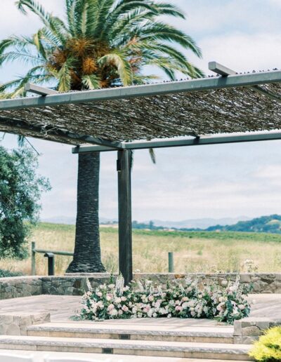 Outdoor wedding altar with a pergola covered in woven branches, adorned with pink and white flowers. Palm and floral surroundings with a scenic countryside backdrop.