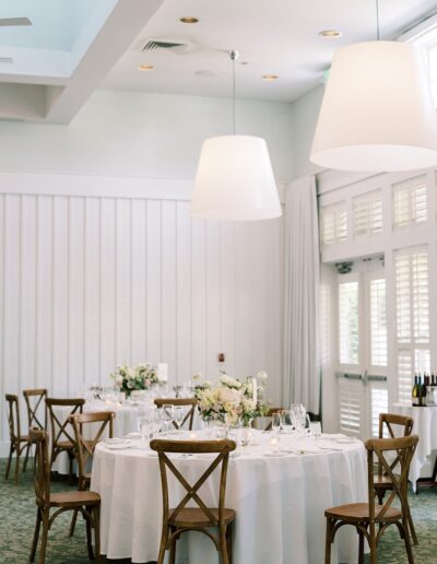 Elegant dining room with round tables set for an event, featuring white tablecloths, floral centerpieces, wooden chairs, and large overhead pendant lights.