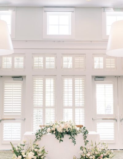 Table decorated with white and pink flowers, set in a bright room with large shuttered windows.