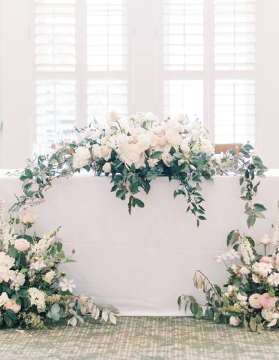 Elegant floral arrangement in white and pastel colors decorates a table covered with a white cloth, set against large windows with shutters.