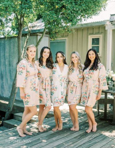 Five women in floral robes stand together on a wooden deck near a tree and a small building, smiling at the camera.