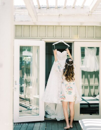 A woman in a floral robe holds a white wedding dress on a hanger in front of double doors on a wooden deck.