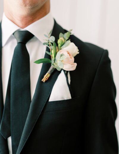 A person in a black suit and black tie, wearing a white shirt. A boutonniere with white and pink flowers is pinned to the lapel.