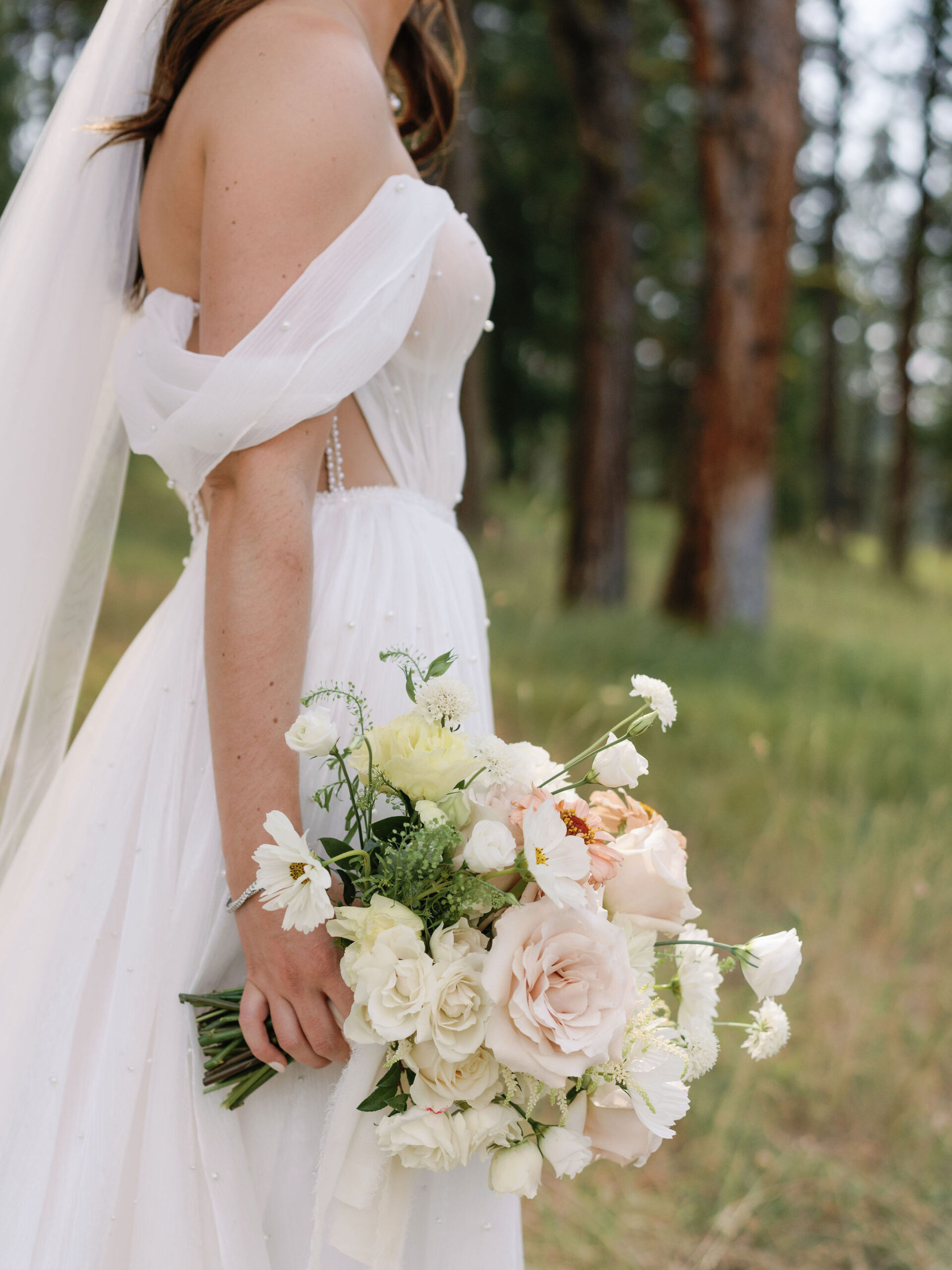 Bride in white strapless gown holds a bouquet of roses and daisies in an outdoor setting.