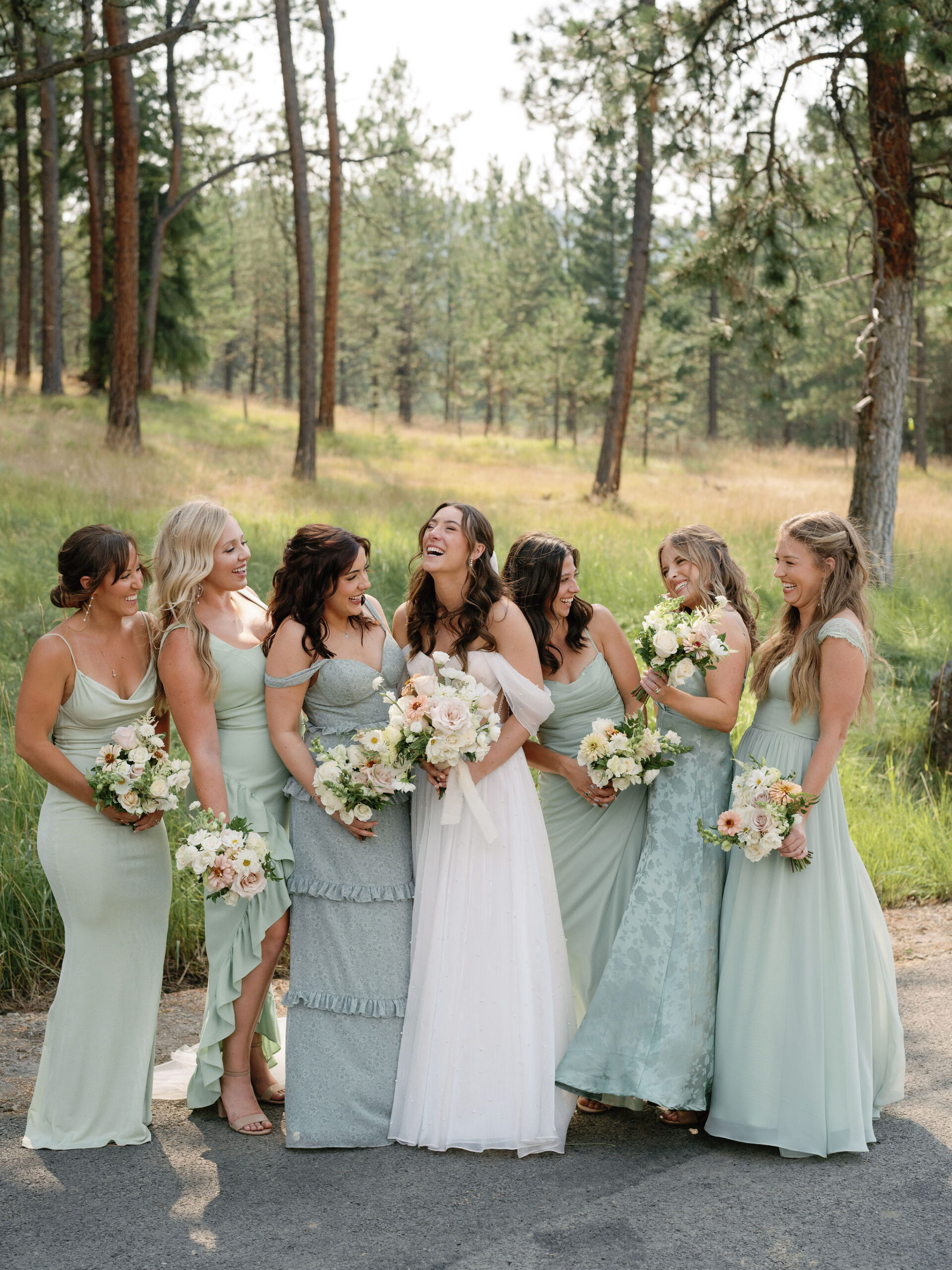 A bride in a white dress stands outdoors with six bridesmaids in light green dresses, all holding bouquets and smiling.