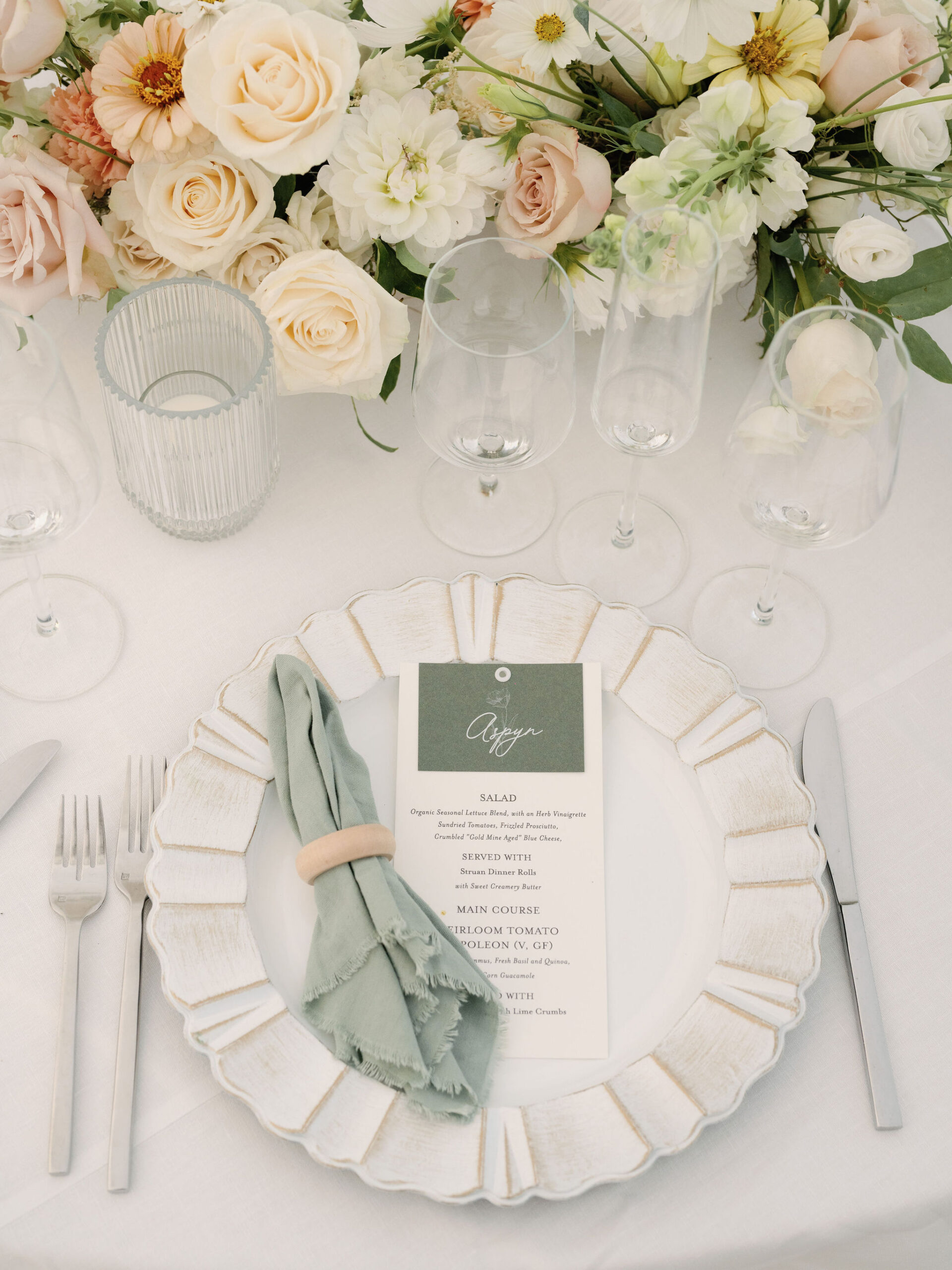 Elegant table setting with a white plate, green napkin, menu card, wine glasses, and a floral centerpiece of white and peach flowers.