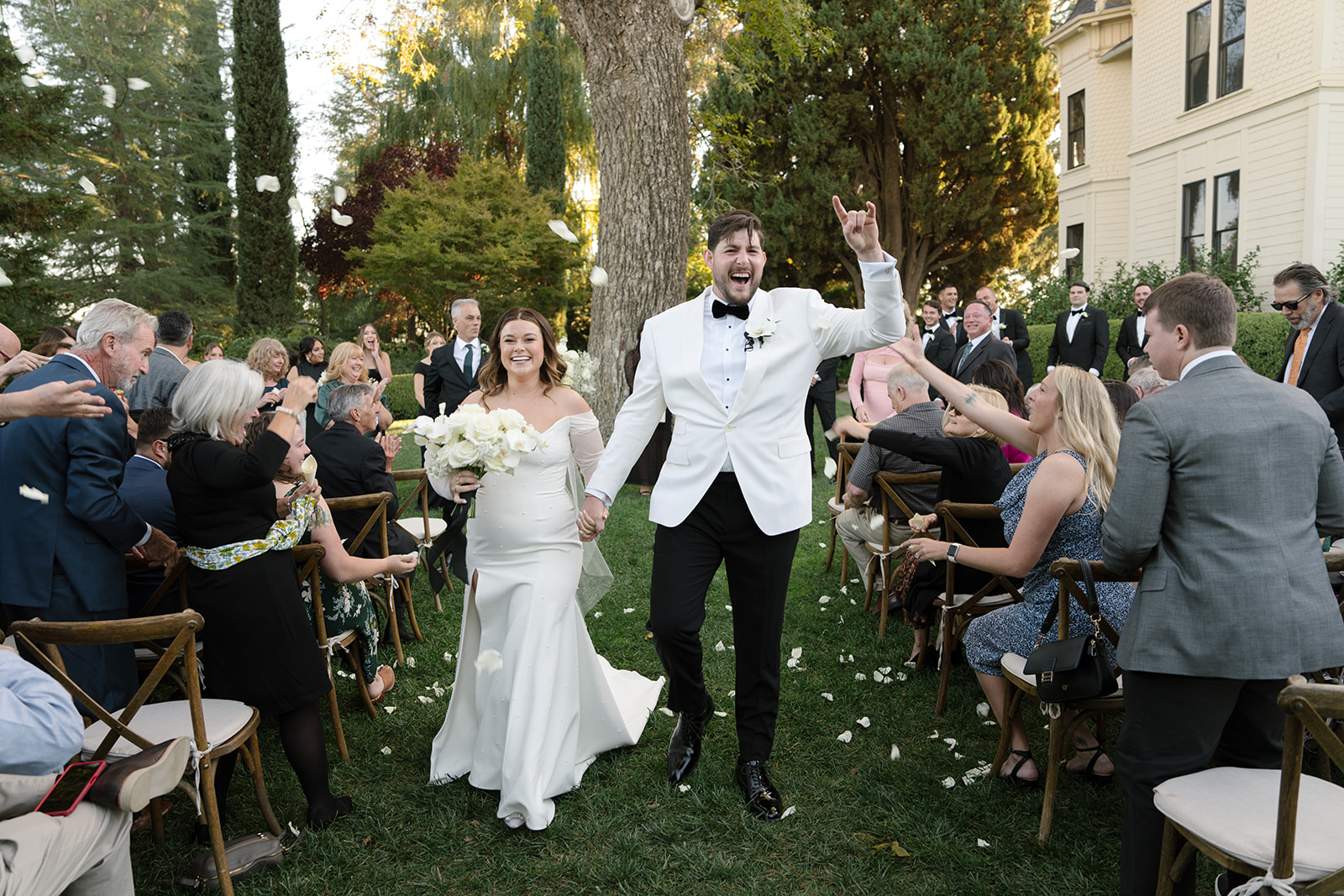 Bride and groom walk down the aisle outdoors, smiling and holding hands, surrounded by seated guests tossing flower petals.