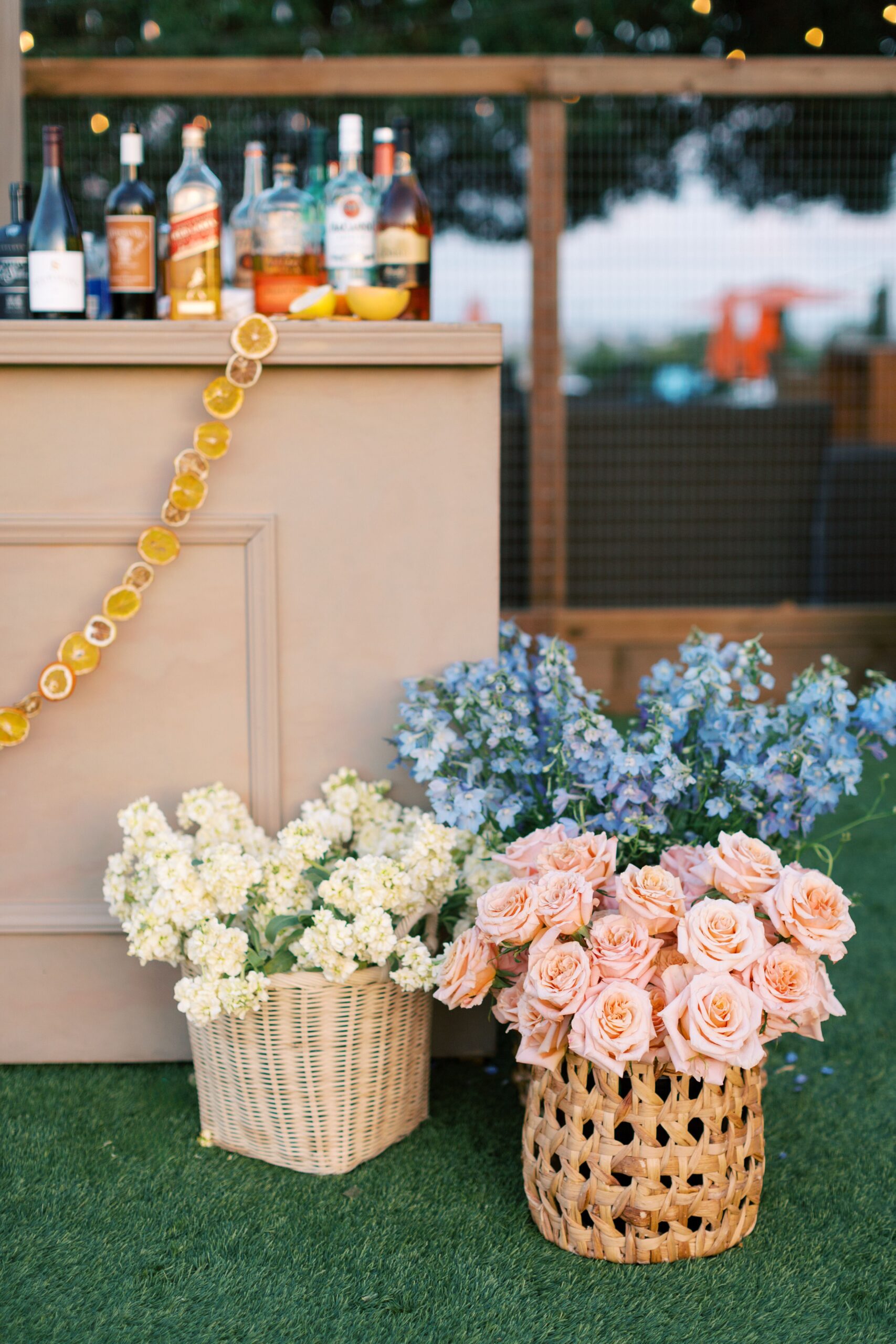 A wooden bar with liquor bottles on top. Two baskets of flowers, one with white blossoms and the other with pink roses, are placed on the green grass beside the bar.