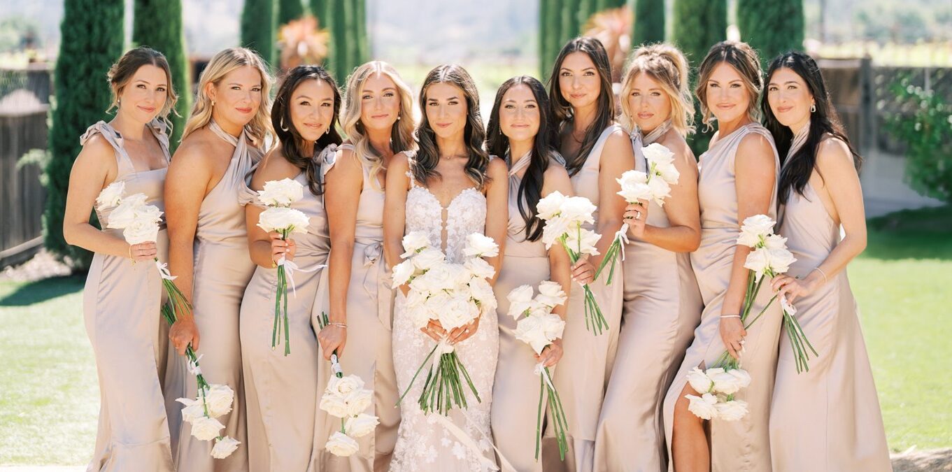 A bride stands with her bridesmaids in a garden setting; they all wear beige dresses and hold white bouquets.