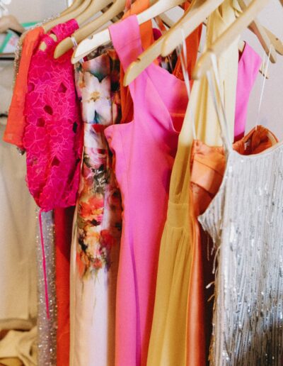 A selection of colorful dresses, including pink, floral, orange, and a silver sequin dress, hanging on wooden hangers in a closet.