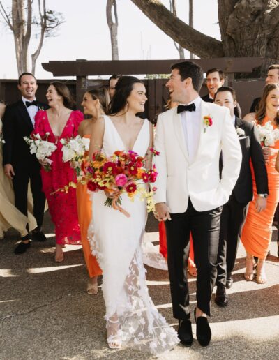 A wedding party walks outdoors, featuring a bride in a white dress and a groom in a white jacket. Bridesmaids and groomsmen follow, wearing colorful dresses and black suits.