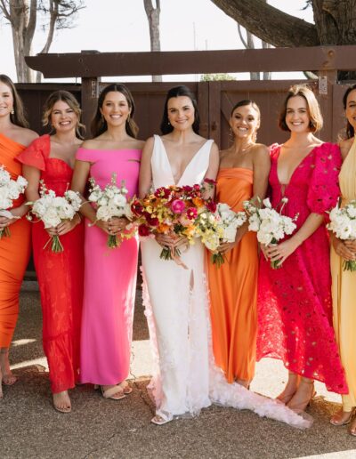 Seven women in colorful dresses stand side by side outdoors, holding bouquets.