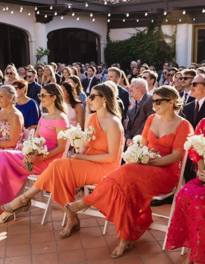 A group of people seated outdoors at a formal event, with women in bright dresses holding white floral bouquets, and string lights overhead.