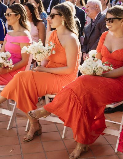 A group of people seated outdoors at a formal event, with women in bright dresses holding white floral bouquets, and string lights overhead.