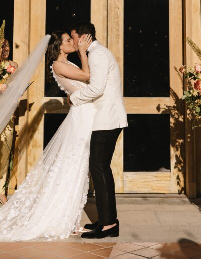 A bride and groom kiss in front of large wooden doors, flanked by floral arrangements with orange and pink flowers.