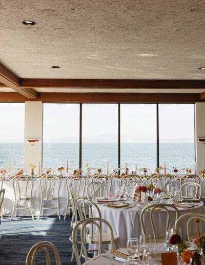 A dining room with round tables set for an event, each with white tablecloths and floral centerpieces. Large windows offer a view of the sea.