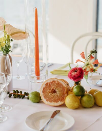 Elegant table setting with fruit centerpiece, flowers, glasses, and an orange candle on a white tablecloth. Scenic view through large windows.