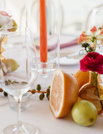 Elegant table setting with empty wine glasses, sliced grapefruit, whole pears, flowers in vases, and a tall orange candle on a white tablecloth.