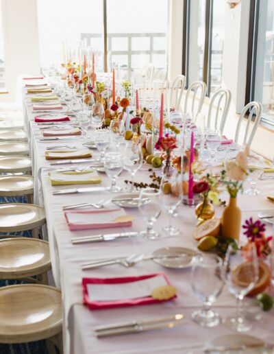 A long, formal dining table set with white tablecloths, arranged with glasses, cutlery, colorful flowers, and candles, surrounded by white chairs.