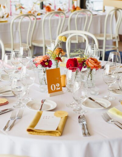 A round table set for a formal event with glasses, cutlery, and colorful napkins. Centerpiece includes red and orange flowers. A table number card marked "02" is displayed.