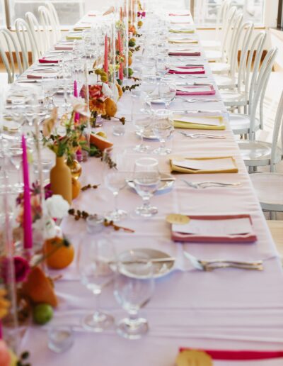 A long dining table is elegantly set with white linens, colorful napkins, candles, and floral centerpieces. Wine glasses and cutlery are neatly arranged at each place setting.