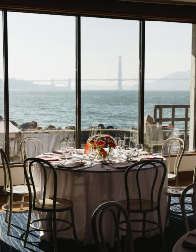 A round dining table set for six by a window with a view of a large bridge over water in the background.