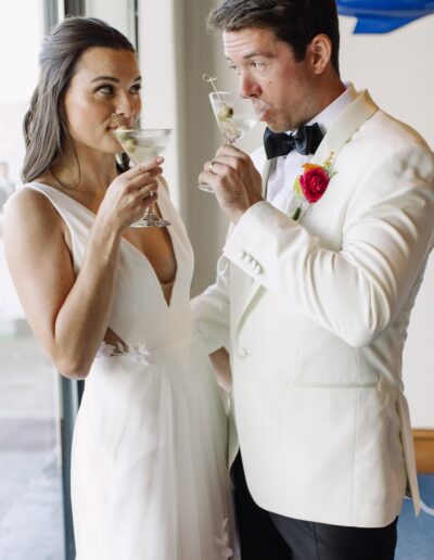 A couple in formal attire, both wearing white, enjoy cocktails with a green olive garnish. The woman looks towards the man as they both raise their glasses.