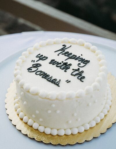 Round white cake with piped icing words reading "Keeping up with the Boness" on top, surrounded by a decorative white border, on a gold and white base.