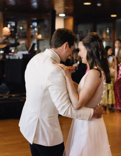 A couple in formal attire dances closely in a warmly lit room with people visible in the background.