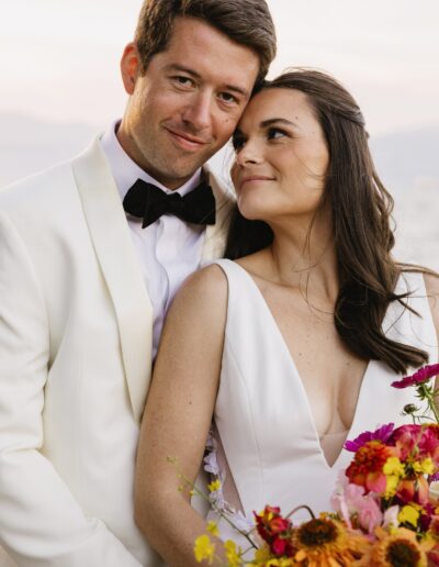 A couple in wedding attire, with the man in a white suit and bow tie, and the woman in a white dress holding a colorful bouquet, stand close with a serene backdrop.