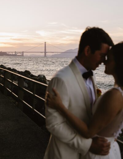 A couple embraces near a railing with a view of the Golden Gate Bridge in the background during sunset.