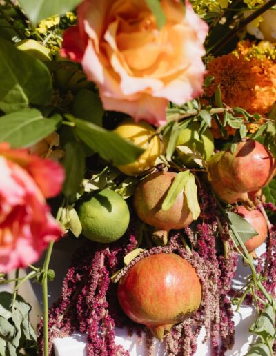 A vibrant floral arrangement featuring roses, marigolds, greenery, pomegranates, and various fruits.