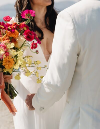 Bride and groom in white attire stand by the water, with the bride holding a colorful bouquet of flowers.