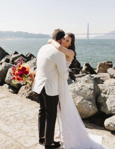 A couple in wedding attire embrace near a rocky shore with a bay and bridge in the background. The bride holds a colorful bouquet.