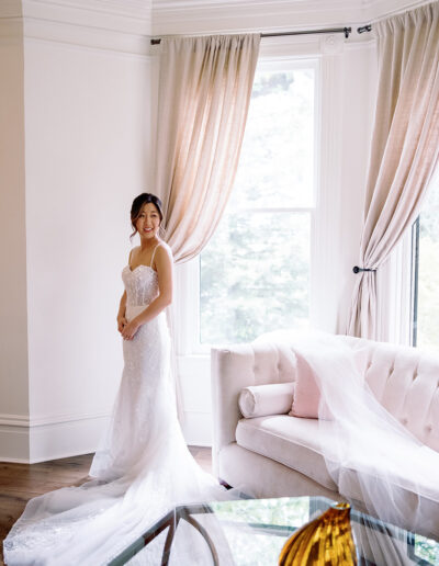 The bride in a lace wedding dress stands by a window in the bright room, reminiscent of the elegant ambiance of Park Winters Weddings, with a sofa and coffee table completing the serene scene.