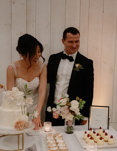 A bride and groom stand by a table with a cake, flowers, and desserts, basking in the charm of Park Winters Weddings, in front of a white wooden wall.