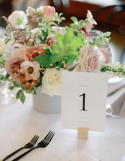 Floral centerpiece with pink and white flowers gracing a Park Winters Weddings table setting, featuring a "Table 1" sign amidst decorative greenery and place settings with forks visible.