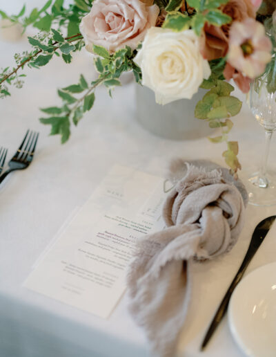 A floral arrangement with pink and white roses graces the table at Park Winters Weddings, accompanied by a menu, a beige napkin, forks, a knife, a plate, and a wine glass.