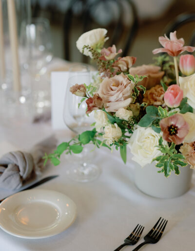 A table setting perfect for Park Winters Weddings, featuring a floral centerpiece with pastel roses and tulips, surrounded by elegant glassware, a white plate, and black cutlery on a crisp white tablecloth.