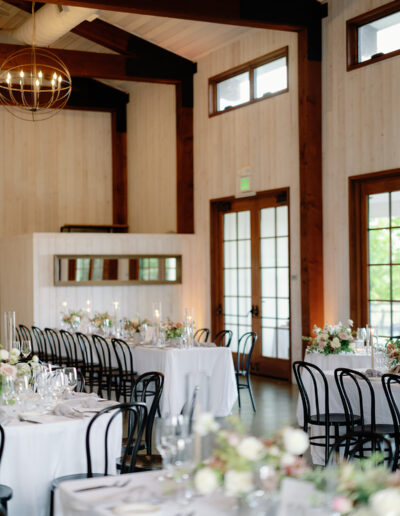 Experience the elegance of Park Winters Weddings with a charming dining setup featuring round tables adorned with white tablecloths, black chairs, and floral centerpieces, all lit by a stunning chandelier in a warm wooden venue.