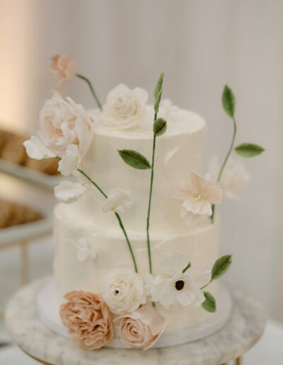 A two-tiered white cake, perfect for Park Winters Weddings, adorned with delicate pink and white flowers and green leaves, rests elegantly on a marble table.