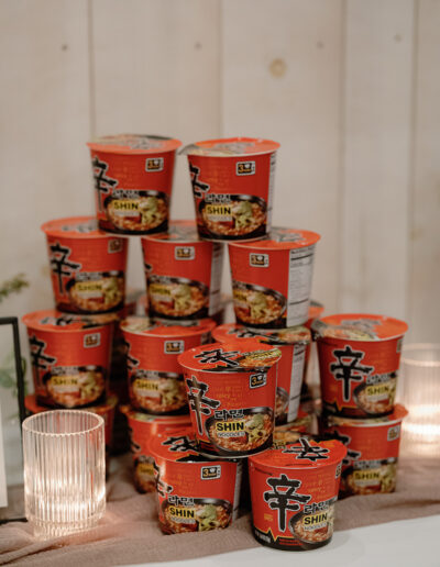 Stacked red containers of Shin Ramyun noodle soup against a light wood background, with candles reminiscent of a Park Winters wedding setting on the sides.