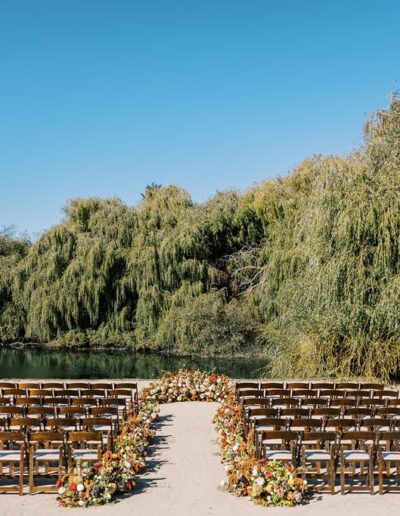 Outdoor wedding setup with rows of wooden chairs arranged along a sandy aisle, decorated with floral arrangements. Surrounded by lush greenery and a pond in the background under a clear blue sky.