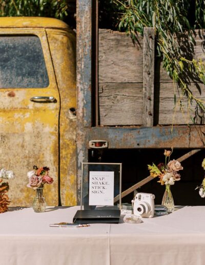 A vintage truck serves as a backdrop for a table with floral arrangements, a sign, a camera, and a guestbook on a white tablecloth.