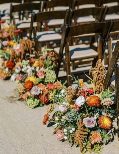 Wooden chairs are lined up in rows on a sandy surface, decorated with colorful floral arrangements including orange, yellow, and pink flowers.